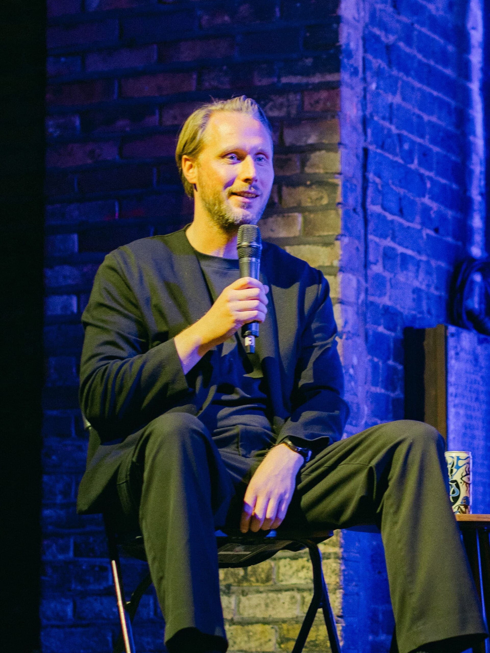 Leo von Hoyningen-Huene, Treasurer of OceanPulse, speaking on stage with a microphone during an event, wearing a dark jacket against a brick wall background with blue lighting.