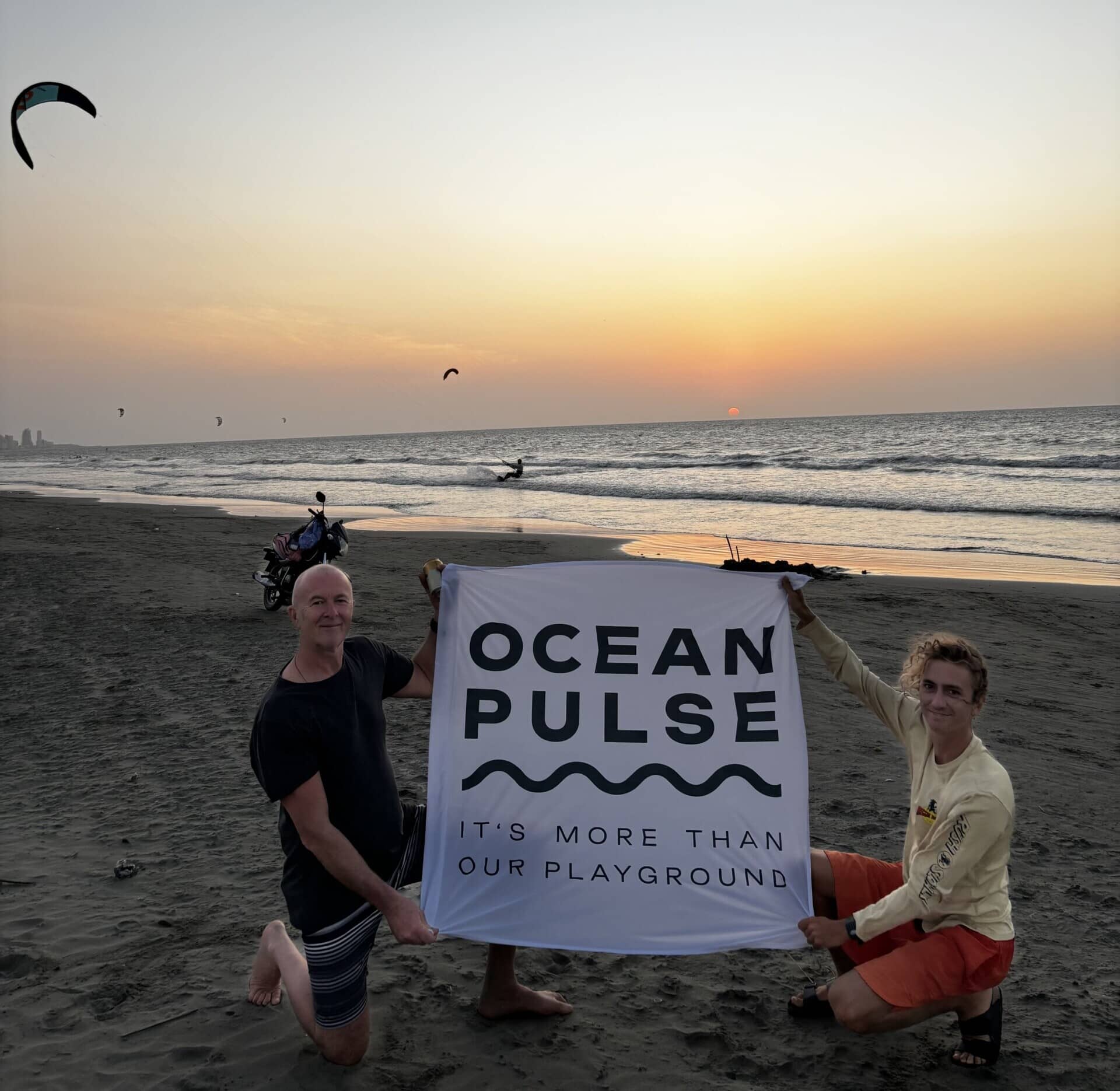 Watersport for Change Instructor Luke Riley und sein Vater Sean halten das OceanPulse-Banner bei Sonnenuntergang am Strand, im Hintergrund sind Kitesurfer zu sehen.