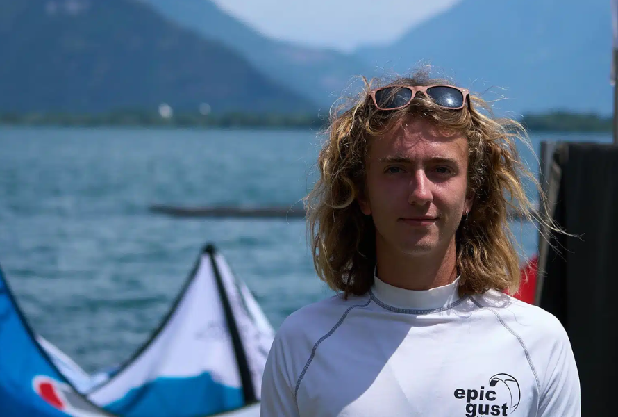 Portrait of Luke Riley, Watersport for Change instructor, standing by the water with kiteboarding kites and mountain scenery in the background.