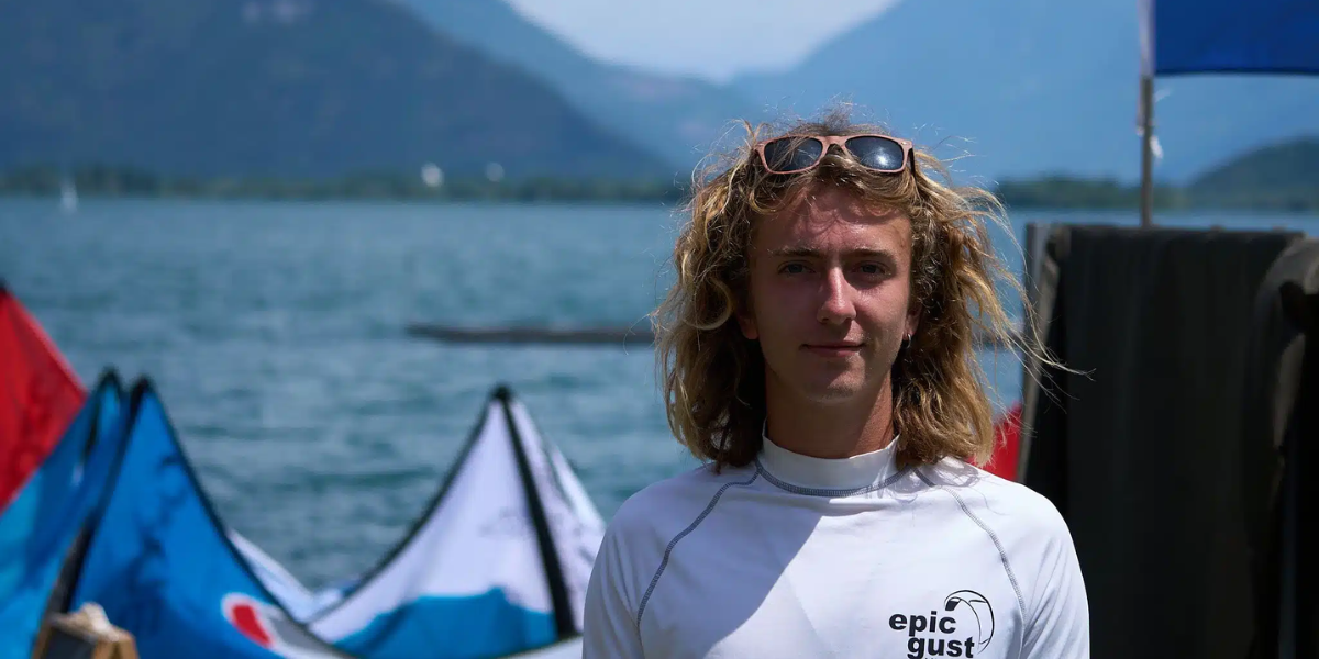 Portrait of Luke Riley, Watersport for Change instructor, standing by the water with kiteboarding kites and mountain scenery in the background.