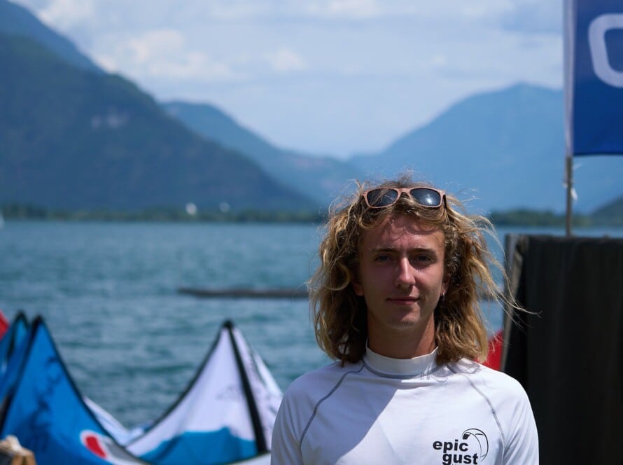 Portrait of Luke Riley, Watersport for Change instructor, standing by the water with kiteboarding kites and mountain scenery in the background.