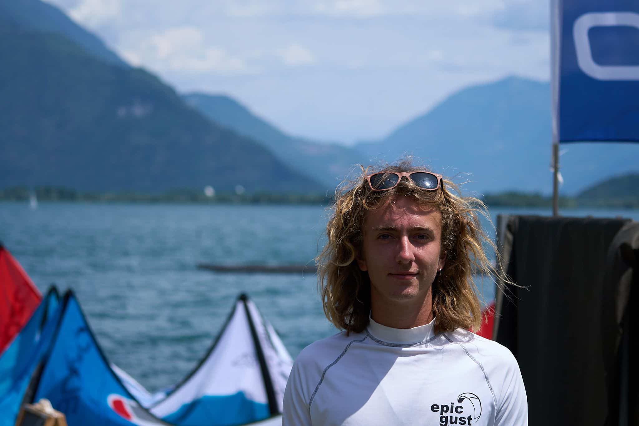 Portrait of Luke Riley, Watersport for Change instructor, standing by the water with kiteboarding kites and mountain scenery in the background.