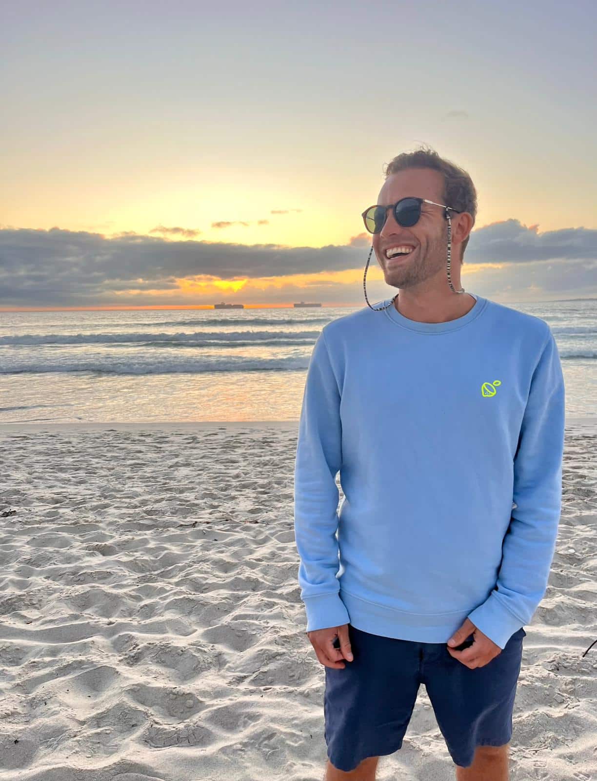 Timo Rogge, Vice-Chairman of OceanPulse, standing on a beach at sunset smiling, wearing a light blue sweatshirt and sunglasses with the ocean and ships in the background.